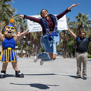 A student jumping up mid air as Sammy Spartan and another person holds a huge scholarship check.