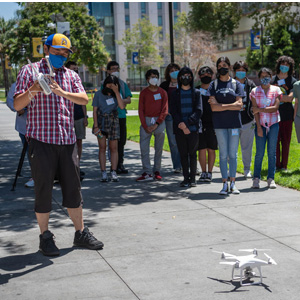 A student holding a remote for their drone that is on the ground and about to take off.
