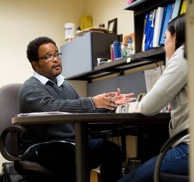 A faculty member advises a student at his desk.