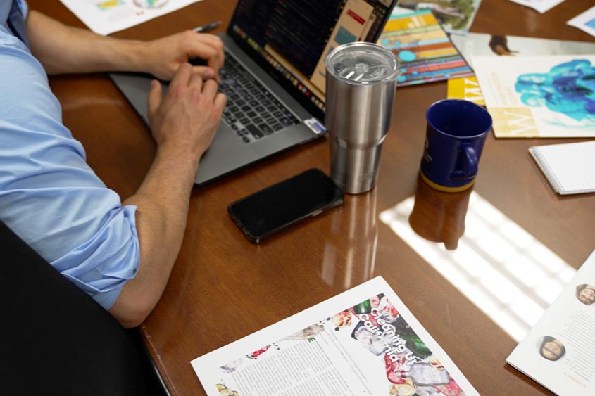 A staff member working on his laptop on a desk with some printed materials spread throughout.