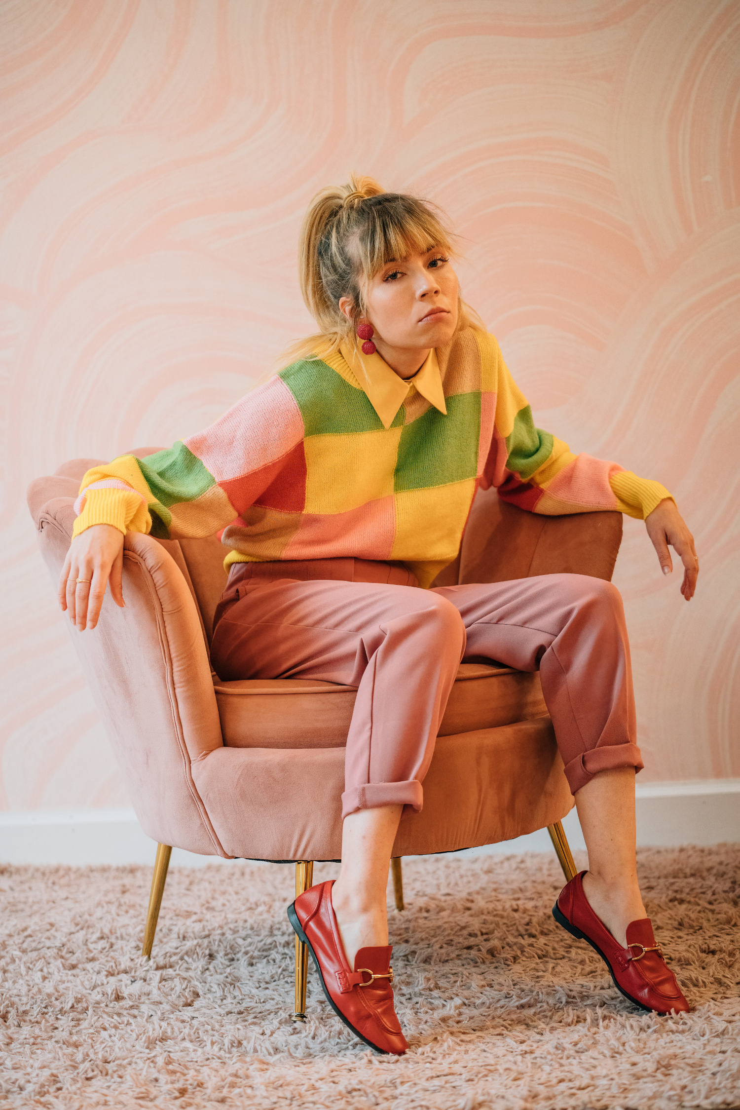 Colorful retro fashion portrait of woman seated in pink chair Woman in colorful sweater and pink pants sitting in a pink chair against a pastel patterned wall.