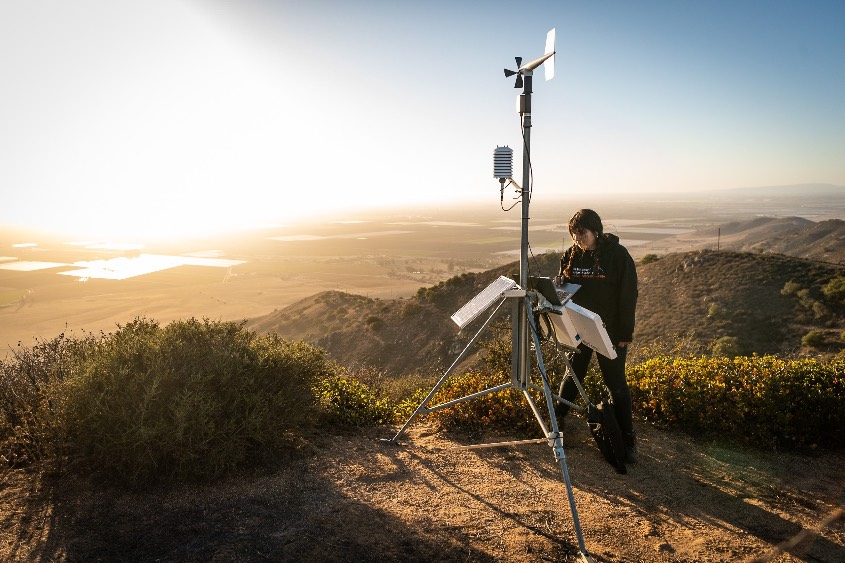 A students stands at the top of a high mountain while working on equipment to track wildfires.