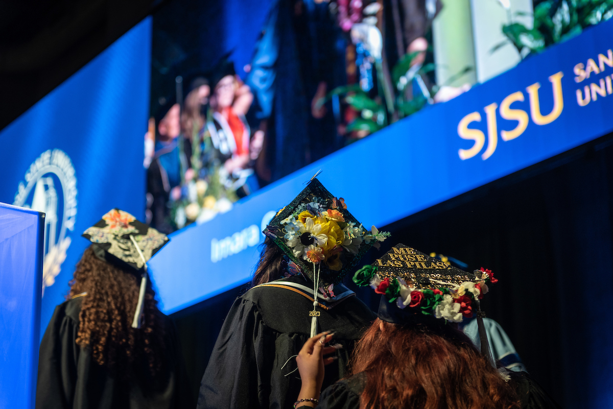 Female student in cap and gown walking up to podium, back facing the camera 