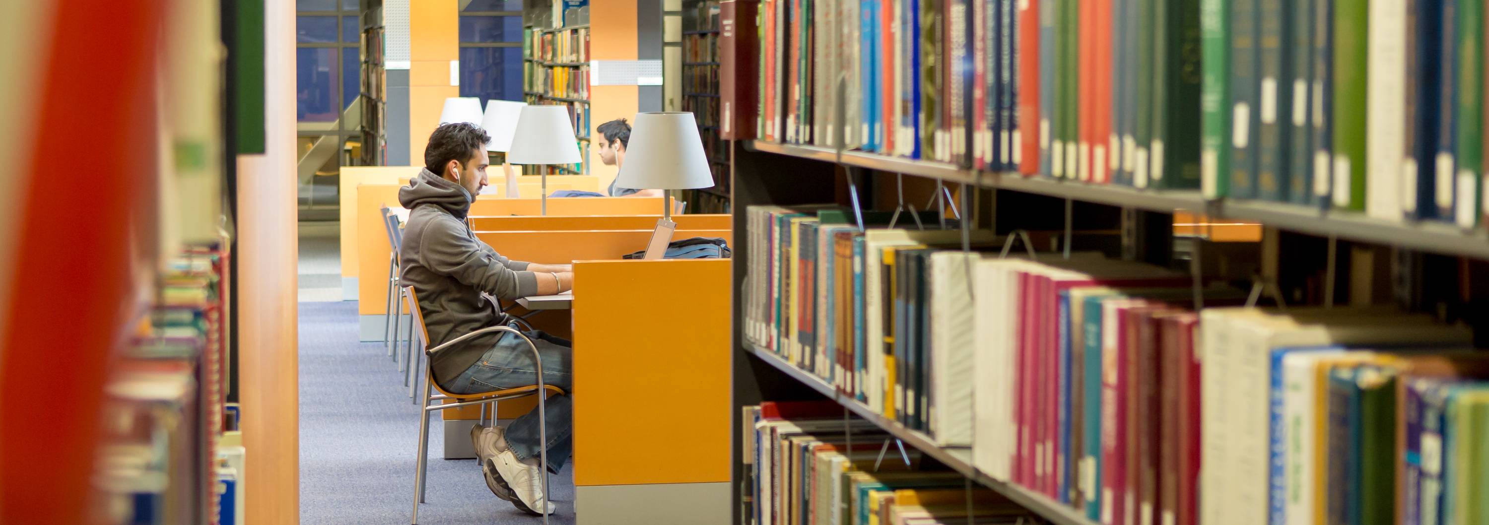 Student sits in a desk in the library, bookcases frames his figure in the chair.