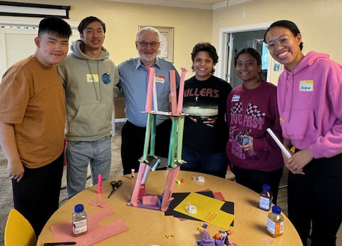 Students pose with a Rotary club visitor in front of a sculpture constructed from tape, multi-colored paper, and other office supplies