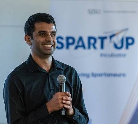 A dark-haired man holds a microphone and smiles, with a white pop-up banner that says "SpartUp Incubator" behind him.