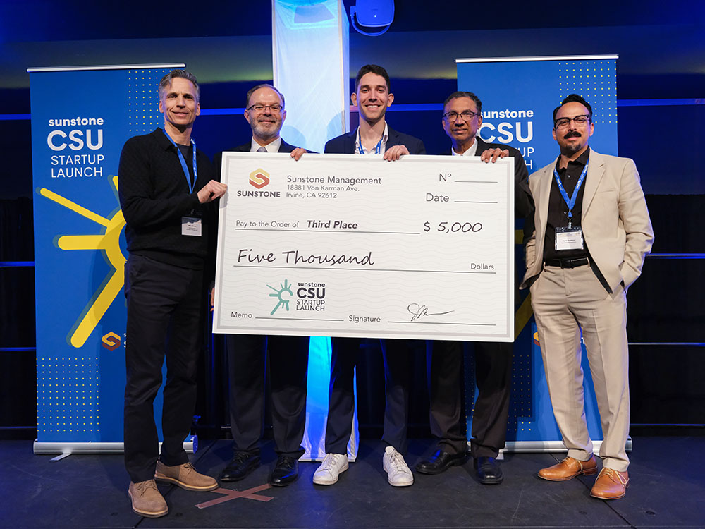 A male, college-age startup founder poses onstage with two sponsors, two hosts, and a giant check for $25,000