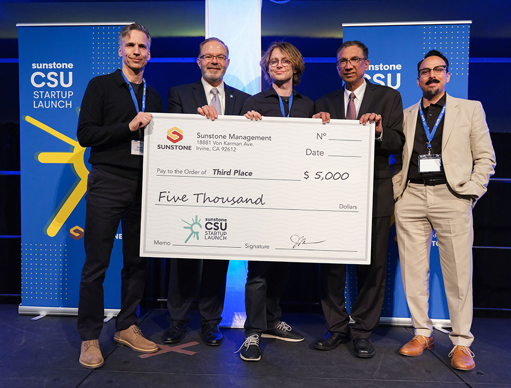 A male, college-age startup founder poses onstage with two sponsors, two hosts, and a giant check for $5,000