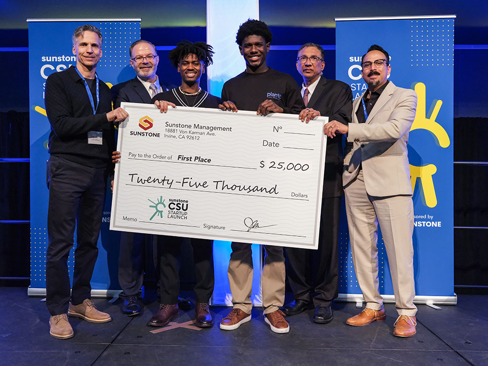 Two male college-age startup founders pose onstage with two sponsors, two hosts, and a giant check for $25,000