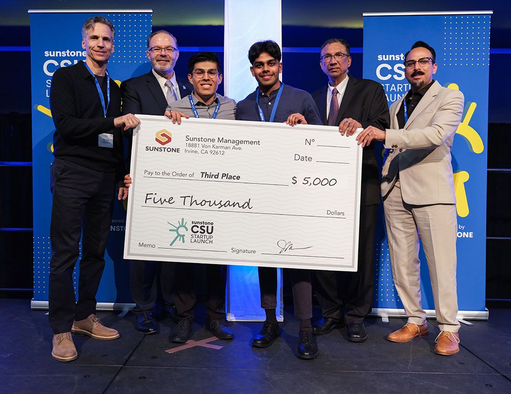 Two college-age startup founders pose onstage with two sponsors, two hosts, and a giant check for $5,000