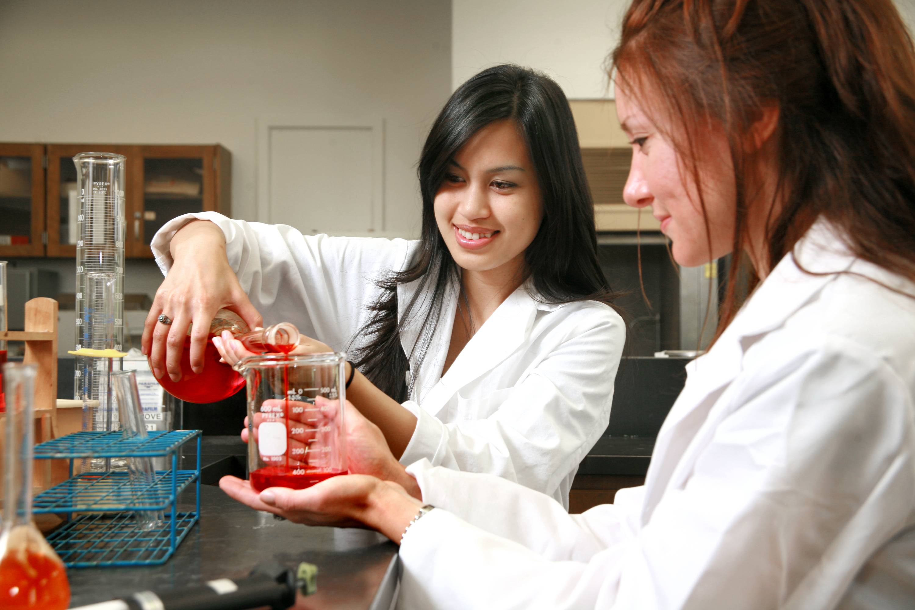 Two female students pour a red liquid from one beaker to another inside a lab.