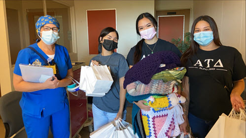 Three Alpha Tau Delta members donating items, including blankets. The three members are darked haired women, wearing masks while obviously smiling. A nurse, dressed in blue, stands and smiles with them.