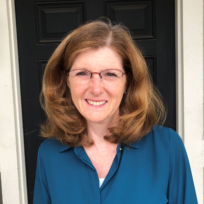 Portrait of Lisa Walker-Vischer, a pale-skinned woman with red hair. She is standing in front of a dark door, and is wearing a blue blouse.