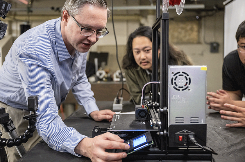 Photograph Prof. Tom Madura demonstrating 3D printer functionality to students.