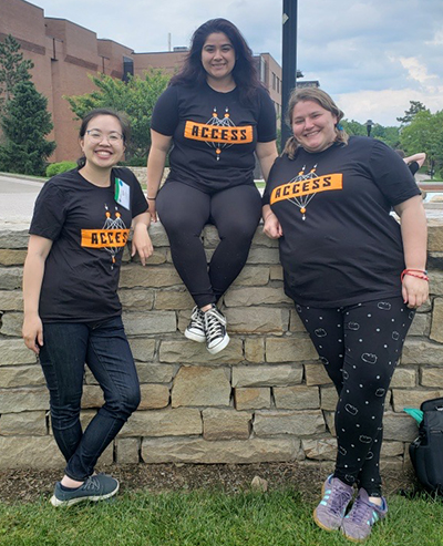 Gina Quan, Autumn Galinski, and Brianne Gutmann pose in front of a strone wall wearing matching Access Network T-shirts.