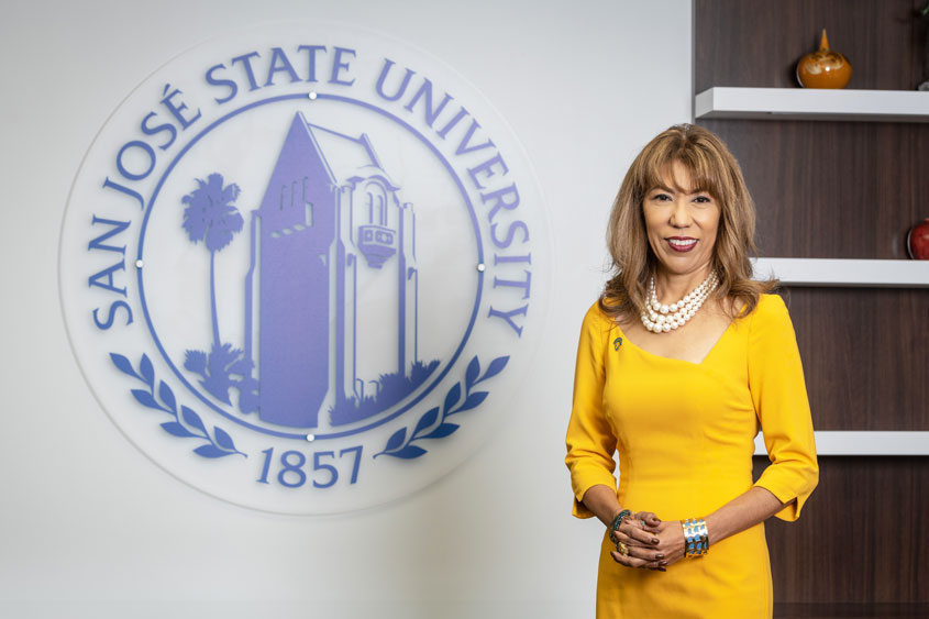 The president in gold dress with white pearls standing in her office with the SJSU seal behind her.