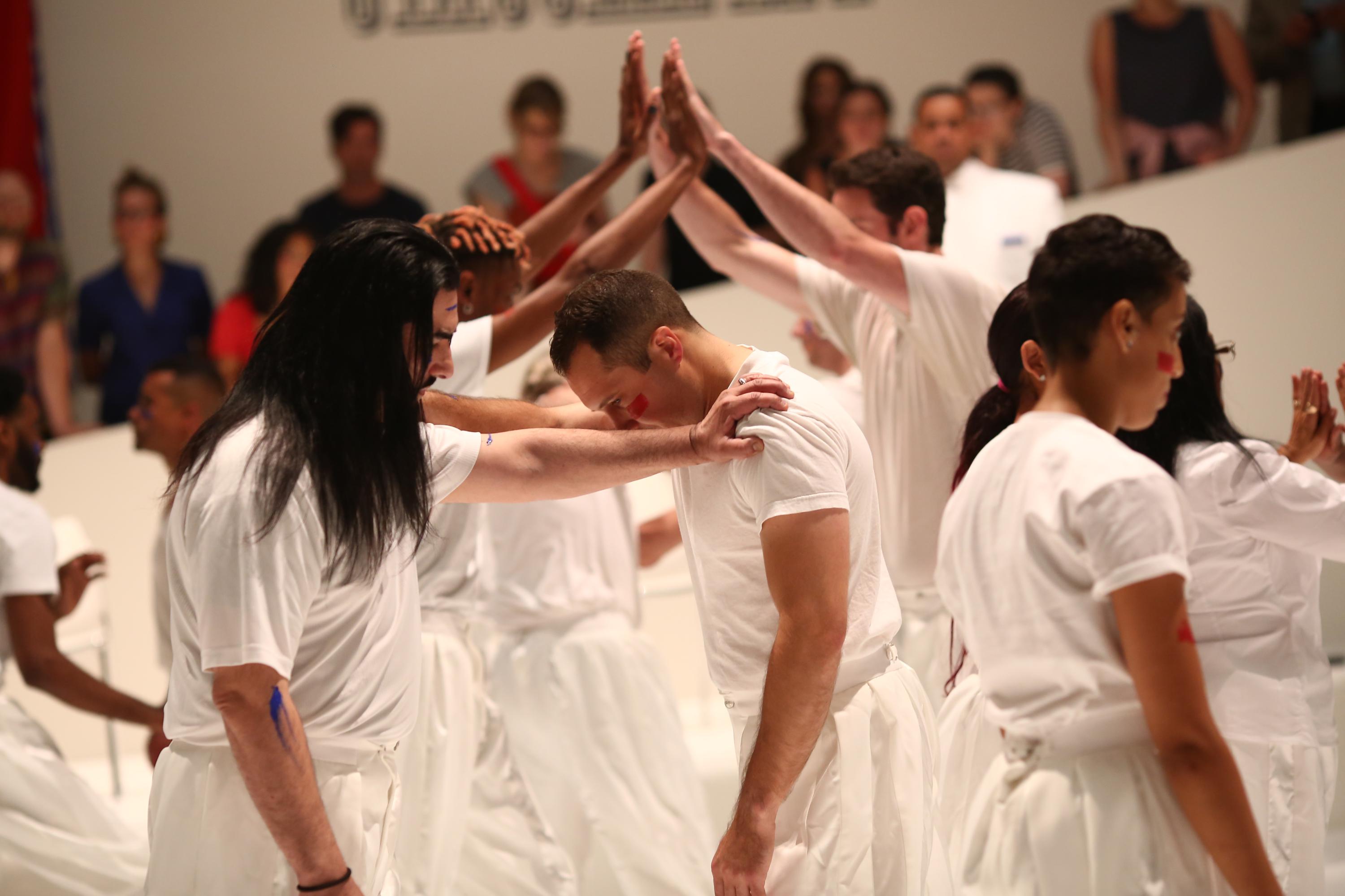 performance still of figures wearing white clothing and joining hands.  The two figures in the foreground rest their hands on each other shoulders