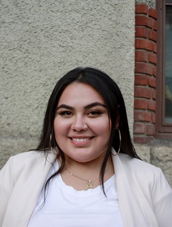 young woman smiling while posing in front of building. She has light medium skin tone, jewelry, white shirt and blazer. 