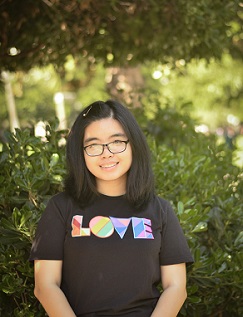 younng woman posing in front of trees and smiling. She has a light medium skin tone, shoulder length hair, and is waring a shirt that reads "love"